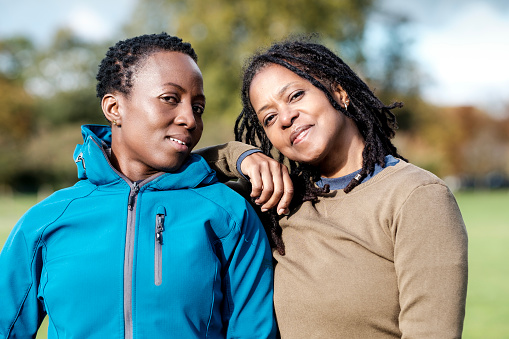 Two mature black women friends posing together in a park in a autumn. It is a cold sunny day and they are wearing warm outfits. One of them is resting her arm on the other one shoulder.
