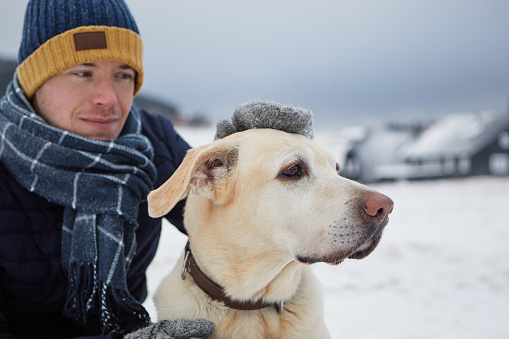 Man dressed in warm jacket, with scarf and cap stroking his dog. Pet owner and labrador retriever against mountain village in winter. Selective focus on dog.