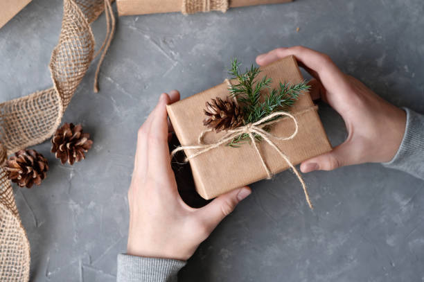 istock Person's hands holding brown crafted gift box decorated with pine branch and cone over gray table background. Sustainable aesthetic Christmas celebration, presents preparing concept 1800314560