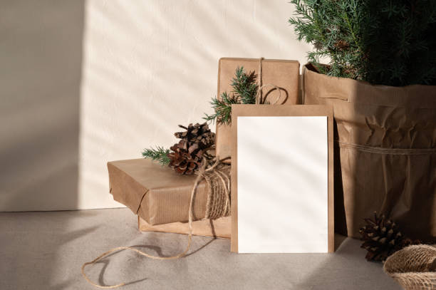 Blank paper card mockup, envelope, crafted gift boxes, juniper tree on neutral beige linen table cloth background with harsh sunlight shadows. Lifestyle Christmas postcard or invitation template