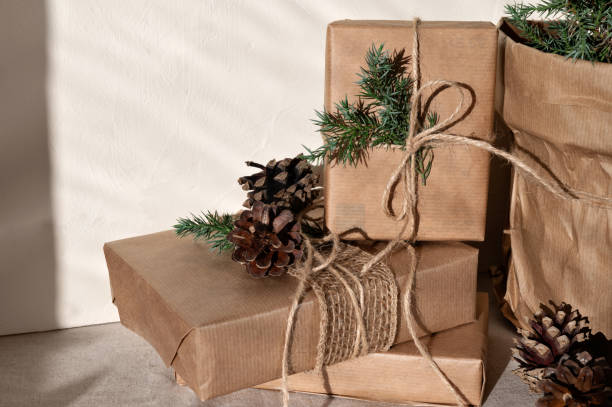 istock Crafted brown gift boxes decorated with pine cones and twigs, on neutral beige linen table background, with harsh sunlight shadows. Lifestyle realistic Christmas composition 1800316663