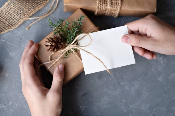 istock Hands holding gift box and blank paper card mockup on gray table background. Christmas sustainable ecological business brand template, craft product holiday package, greeting gift tag or invitation 1800317182