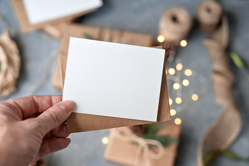 Hand holding blank paper card mockup and envelope over blurred table background with Christmas crafted gift boxes and sparkling garland lights. Christmas greeting postcard or invitation template.