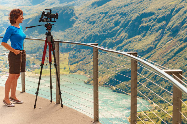 Female tourist with camera on tripod taking travel picture, enjoying mountains landscape from Ornesvingen viewing platform, Norway.