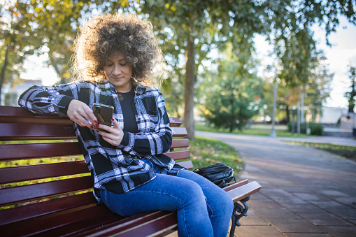 Beautiful young smiling woman with afro hairstyle in casual clothes is sitting on the bench at the public park and using her mobile phone. Wireless communication, social media, blogging, shopping online, banking anywhere.