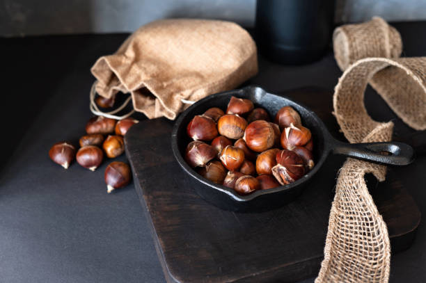 istock Country kitchen table with black wooden board, pan with roasted edible chestnuts. Authentic winter traditional snack cooking 1800399209