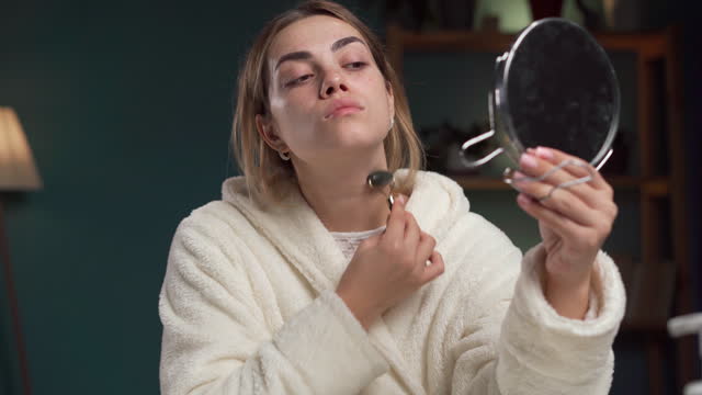 Beautiful woman wearing white bathrobe after shower, making face massage using a jade face roller in her bedroom