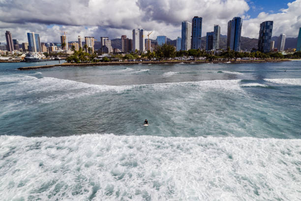 A wide, aerial view of an unrecognizable surfer sitting on their surfboard in the ocean, with the city of Honolulu, Hawaii in the background.