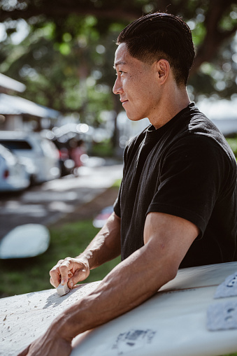 Cropped view of a fit multiracial man of Hawaiian and Japanese descent smiling as he waxes his surfboard near the parking lot of a beach park in Hawaii before heading out to the ocean to surf.