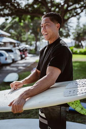 A fit multiracial man of Hawaiian and Japanese descent smiles as he waxes his surfboard near the parking lot of a beach park in Hawaii before heading out to the ocean to surf.
