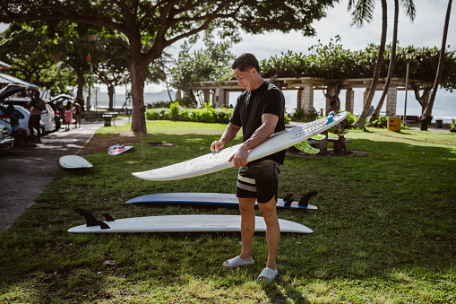 A multiracial young man of Hawaiian and Japanese descent smiles while waxing his surfboard in the grass at a beach park in Hawaii before heading out to the ocean.