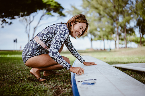 An active and beautiful mature adult Eurasian woman of Hawaiian and Finnish descent smiles while waxing her surfboard in the grass at a public beach park in Hawaii on a beautiful, sunny day.