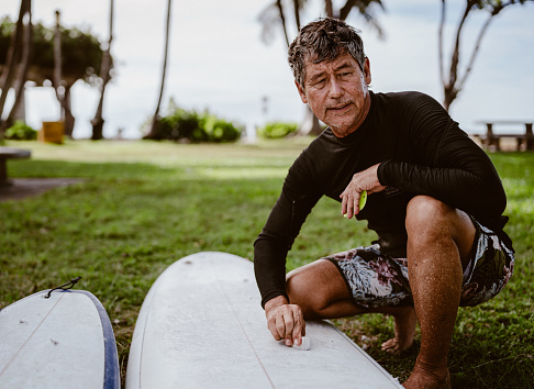 An active and healthy senior male of Hawaiian and Finnish descent waxes his surfboard in a grassy beach park in Hawaii before going surfing.