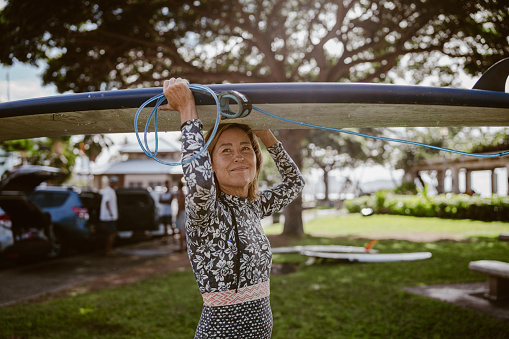 A beautiful and fit mature adult Eurasian woman of Hawaiian and Finnish descent smiles and carries her surfboard above her head as she walks through a grassy public beach park toward the ocean.