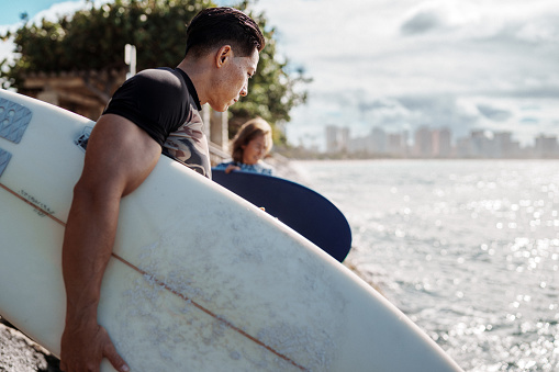 Profile view of a young and fit male surfer of Hawaiian and Japanese descent navigating a rocky shoreline while carrying his surfboard to the ocean with a view of Honolulu, Hawaii visible in the background.