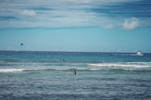 A wide view of an unrecognizable woman sitting on a surfboard in the Pacific Ocean off the coast of Hawaii with a boat and tourists parasailing in the background.
