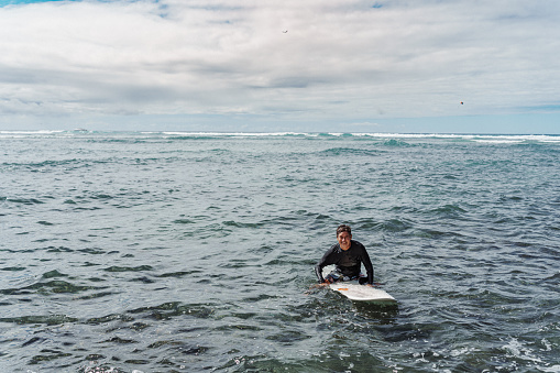 An active Eurasian senior man adult man of Hawaiian and Finnish descent sits on his surfboard and smiles directly at the camera while waiting for the prefect wave off the coast of Hawaii.
