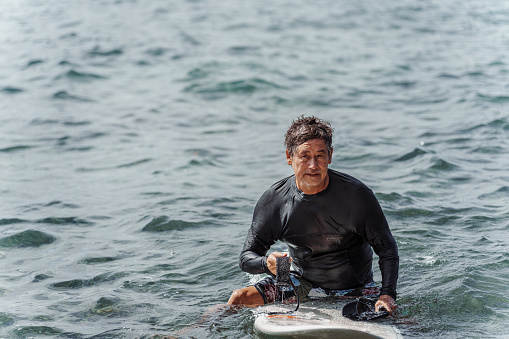 A fit Eurasian senior man of Hawaiian and Finnish descent wearing a wetsuit sits on his surfboard and floats in the ocean off the coast of Hawaii.