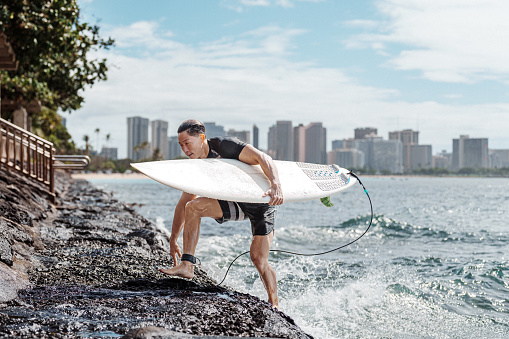 A handsome and fit man of Hawaiian and Japanese descent holds his surfboard and climbs out of the ocean onto the rocky shoreline after surfing in Hawaii with the Honolulu skyline visible in the background.