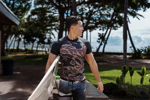 A young man of Hawaiian and Japanese ethnicity looks to his side as he carries his surfboard through a public beach park in Hawaii after spending the morning surfing.