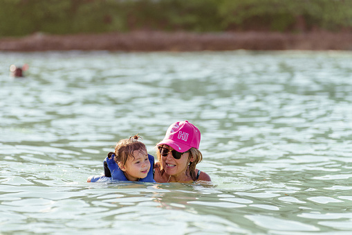 An active Eurasian senior woman helps her three year old granddaughter, who is wearing a life jacket, swim in the ocean while on a family vacation in Hawaii.