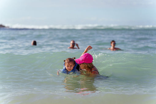 An active Eurasian senior woman helps her three year old granddaughter, who is wearing a life jacket, swim in the ocean while on a family vacation in Hawaii.