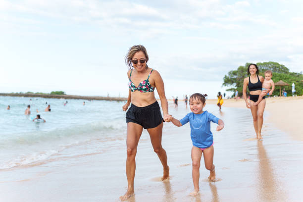 Loving grandmother playing at the beach with her happy granddaughter A beautiful and active senior Eurasian woman holds hands with her three year old granddaughter and playfully runs along the sandy shoreline while at the beach in Hawaii with her adult daughter and grandchildren. family beach vacations travel stock pictures, royalty-free photos & images