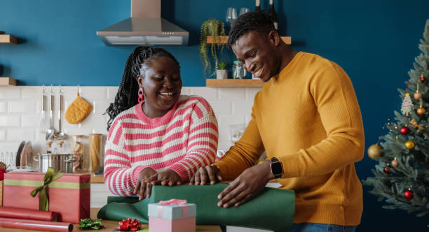 Beautiful young African couple looking happy while wrapping Christmas gifts in colorful paper at home