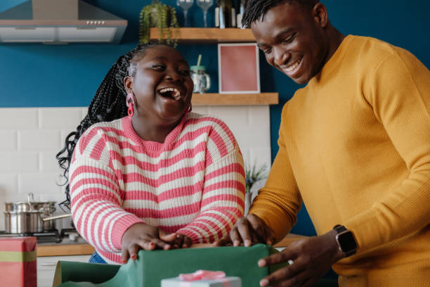 Beautiful young African couple laughing while wrapping Christmas gifts in colorful paper at home together