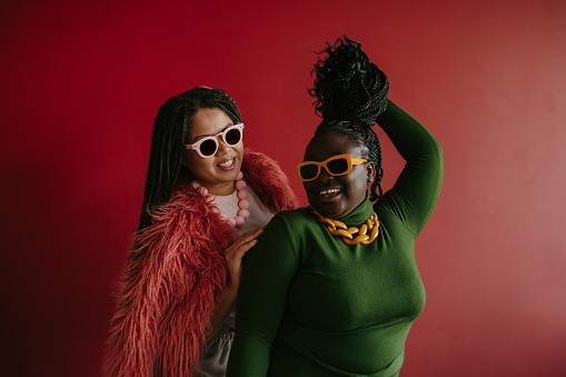 Two joyful plus size African woman playing with their braided hair while having fun on red background