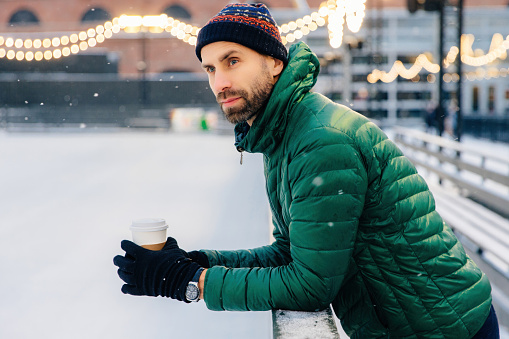 Contemplative man with a coffee cup at a snowy ice rink, festive lights above