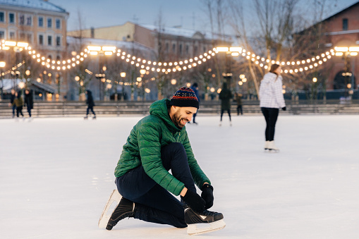 Man tying his ice skates on a festive lit rink at dusk, with skaters behind.