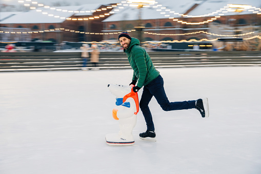 Young male begginer learns skating with help of special figure, leans at it, being on ice rink covered with snow, has cheerful expression, poses in camera. Man uses special equipment for skating