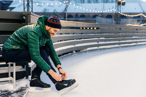 Focused man lacing up ice skates at a snowy rink, with warm lights in background