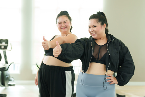 chubby Asian women, standing together in fitness center doing weight loss exercises for sedentary health, women are friends often invite each other come, raise hand make excellent hand signal.