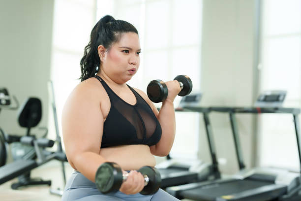 istock Chubby Asian girl Perform a seated exercise by lifting dumbbells with both arms. trying to lose weight serious expression Committed to exercising Overweight Asian woman exercising in the gym. 1800449913