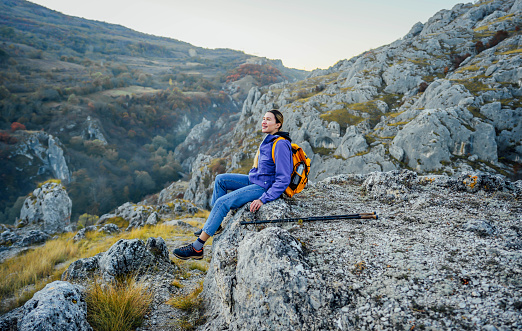 Young smiling woman hiker sitting on mountain ridge and enjoying the view