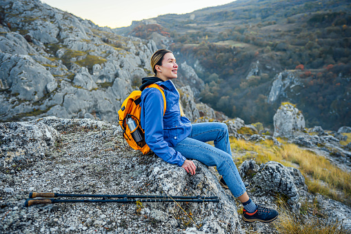Young smiling woman hiker sitting on mountain ridge and looking at view