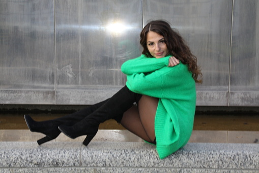 Portrait of an attractive pretty woman in a green mini dress with overknee boots, sitting in front of a silver background