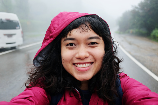 Smiling young Asian woman is travel and selfie in tropical rain forest and raining with fog on the road on vacation in Thailand