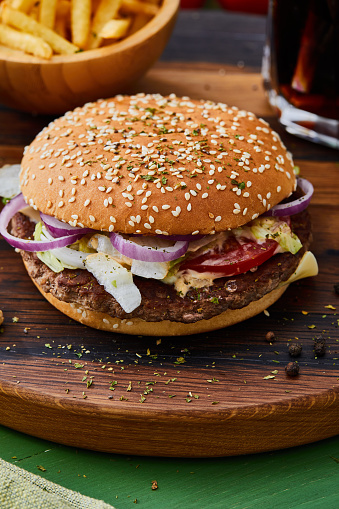 Tasty and delicious beef burger / hamburger / cheeseburger with French fries, cherry tomato, onion sauce, salad and Spanish onion, served  on a wooden cutting board, on bar, restaurant or home green wooden kitchen table background, close up view with copy space, representing fast food and city life, indulgence and joy healthy lifestyle, street food gourmet