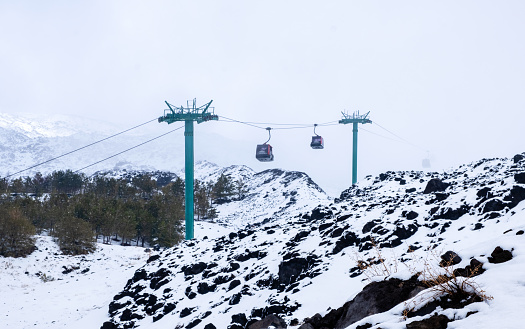 Cable car to Mount Etna national park in winter. View of volcanic terrain with black volcanic lava stones and snow under fog and smoke.