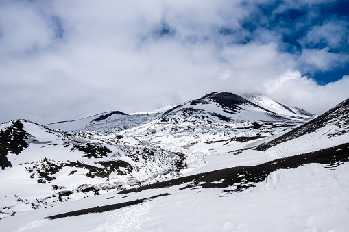 Crater of Mount Etna volcano in winter, Sicily island, Italy. Landscape of Silvestri craters with black volcanic lava stones. Active volcano slopes covered with snow under blue sky, clouds and smoke.