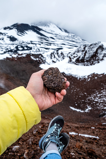 Tourist hand holding lava stone on Mount Etna crater, Sicily, Italy. Warm volcanic lava rock formation. Landscape of craters slopes covered with snow, clouds and smoke.