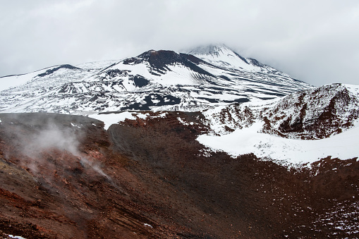 Crater of Mount Etna volcano in winter, Sicily island, Italy. Landscape of Silvestri craters with black volcanic lava stones. Active volcano slopes covered with snow under blue sky, clouds and smoke.