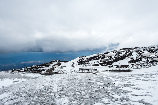 Mount Etna national park in winter. View from volcano crater with black volcanic lava stones and snow under cloudy sky and smoke. View over Sicily island, Italy.