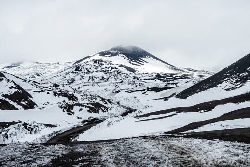Crater of Mount Etna volcano in winter, Sicily island, Italy. Landscape of Silvestri craters with black volcanic lava stones. Active volcano slopes covered with snow under cloudy sky and smoke.