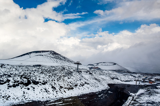 Craters of Mount Etna volcano in winter, Sicily island, Italy. Landscape with black volcanic lava stones, cable car, road and tourist buildings. Slopes covered with snow, blue sky, clouds and smoke.