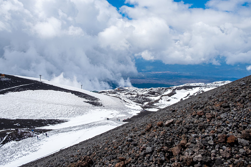 Mount Etna national park in winter. View from volcano crater with black volcanic lava stones, tourists on road and snow under blue sky, clouds and smoke. View over Sicily island, Italy.