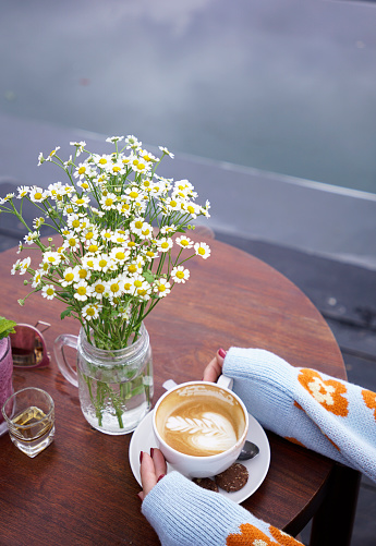 Aerial view of woman in sweater holding a cup of cafe latte on a table decorated with daisies